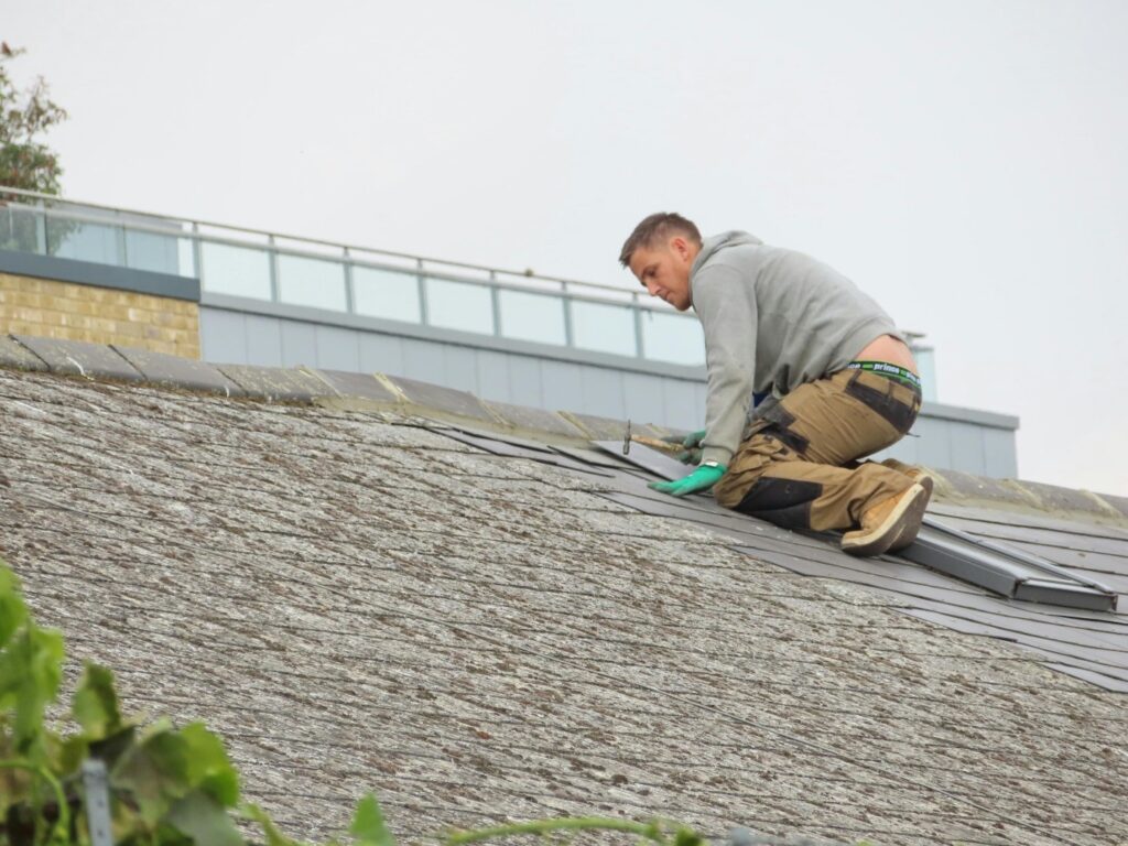 A roofer wearing a gray hoodie, brown work pants, and gloves is repairing a shingle roof